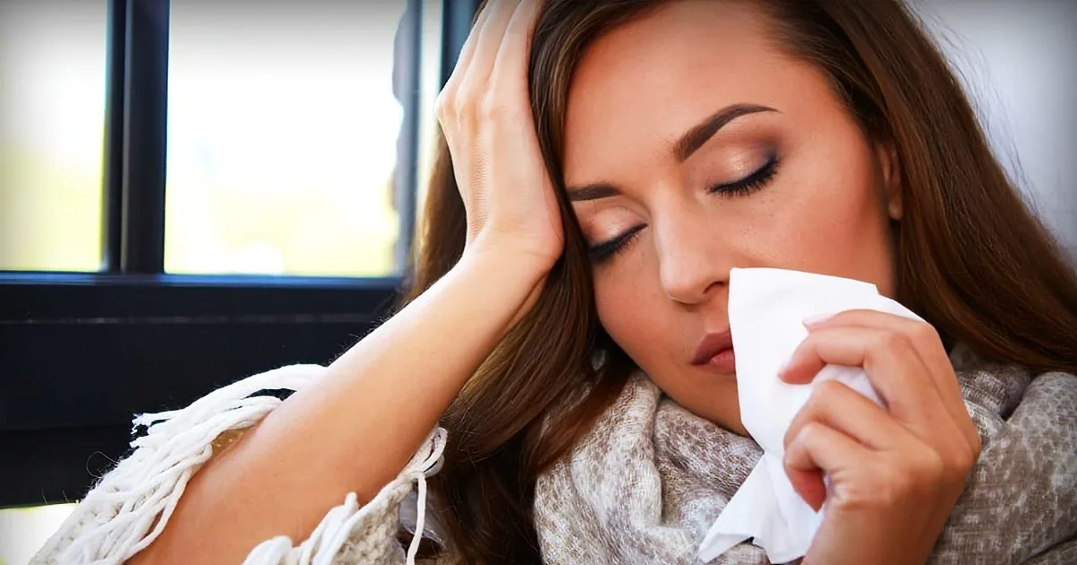 A woman with long brown hair is holding a tissue to her nose, appearing to be sneezing or wiping her nose.