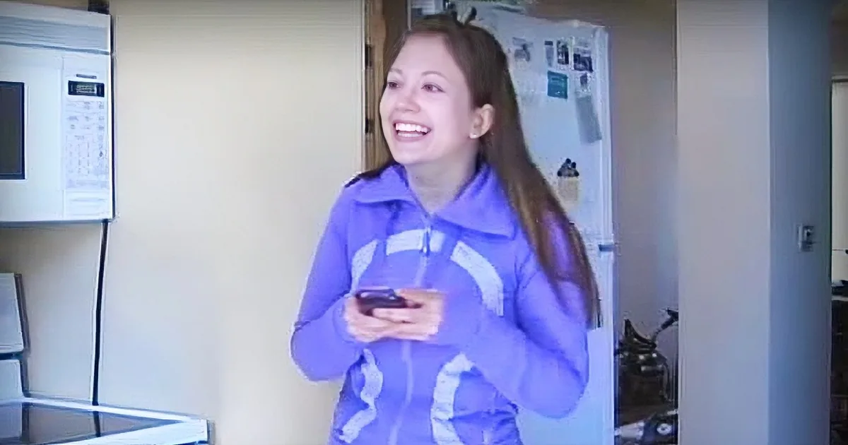 A person in a purple tracksuit holds a phone, smiling, in a kitchen with a microwave and refrigerator.