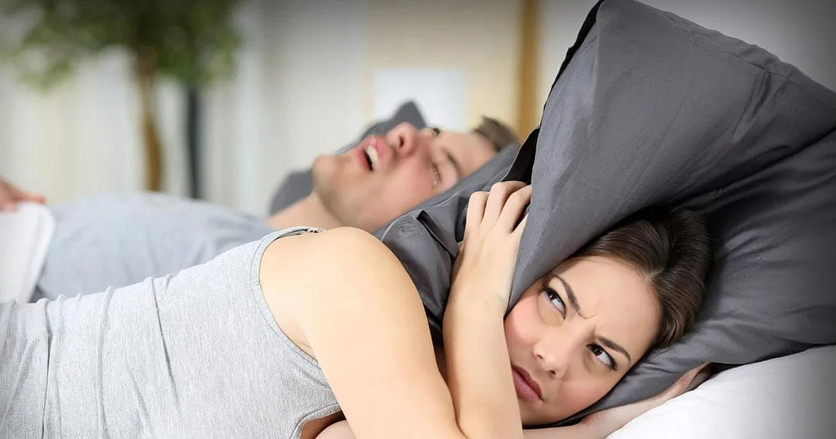 A couple in bed, woman covering her ears while man snores, both in grey.