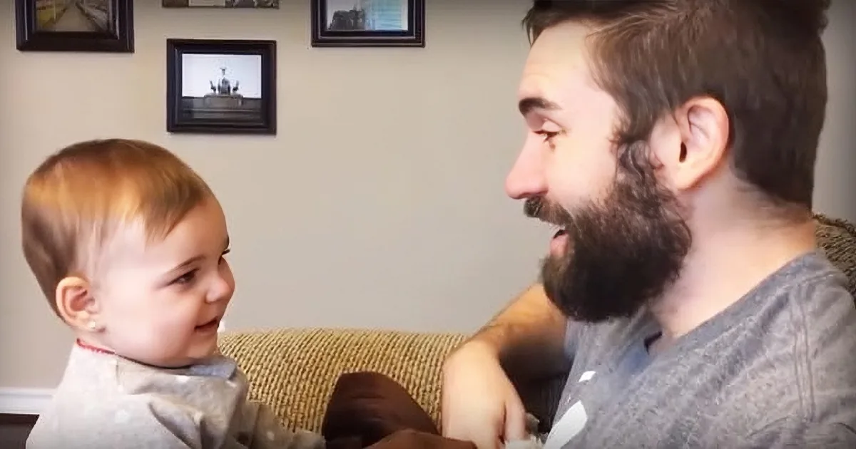 A man with a beard and a baby are smiling at each other, sitting on a couch.