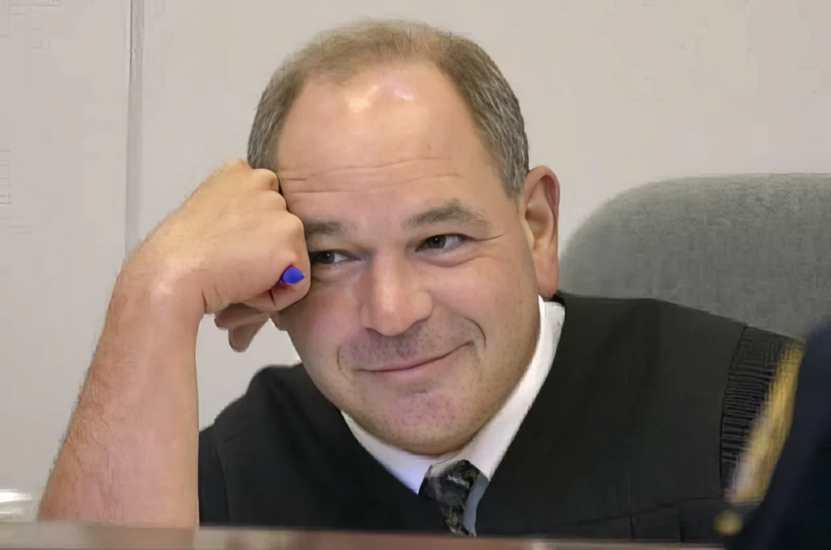 A man in a suit and tie, with his hand on his forehead, smiling slightly.