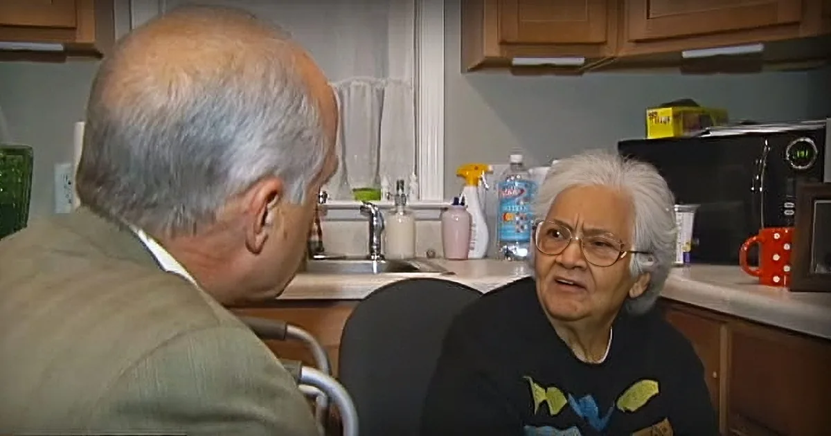 An older man and woman are in a kitchen, with the man speaking and the woman listening attentively.