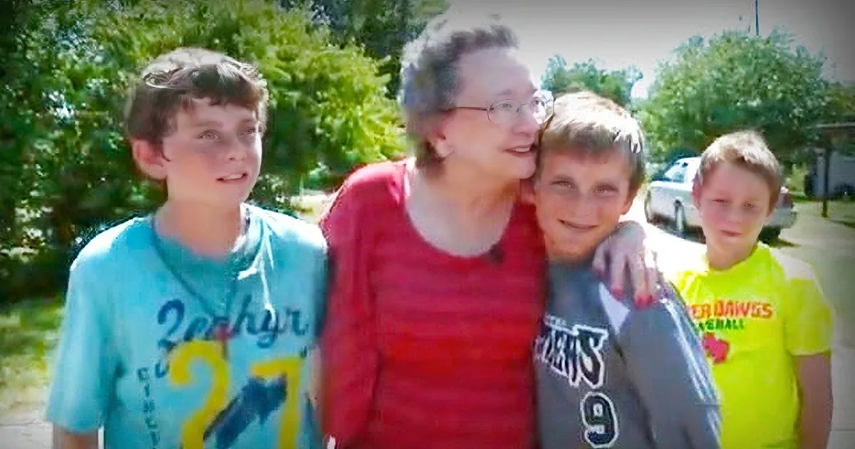 An older woman in red hugs a boy in a yellow shirt, surrounded by two other boys in a sunny outdoor setting.