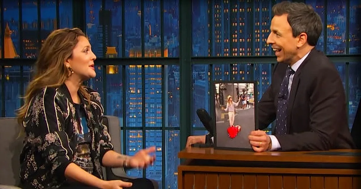 A woman in a floral dress talks to a man in a suit on a talk show set with a cityscape background.