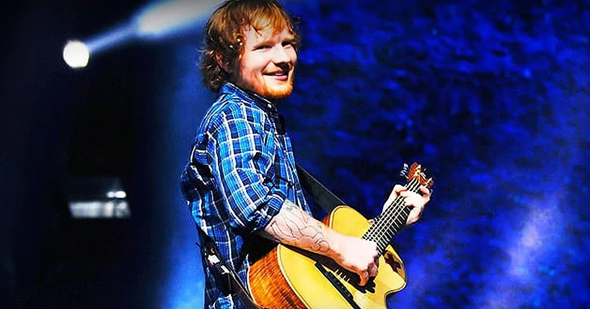 A smiling man in a plaid shirt plays an acoustic guitar on stage with blue lighting.