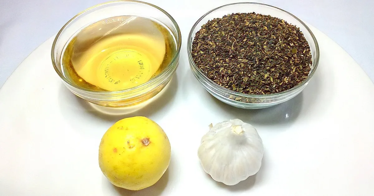 A lemon, garlic, olive oil, and dried herbs are arranged on a white surface.