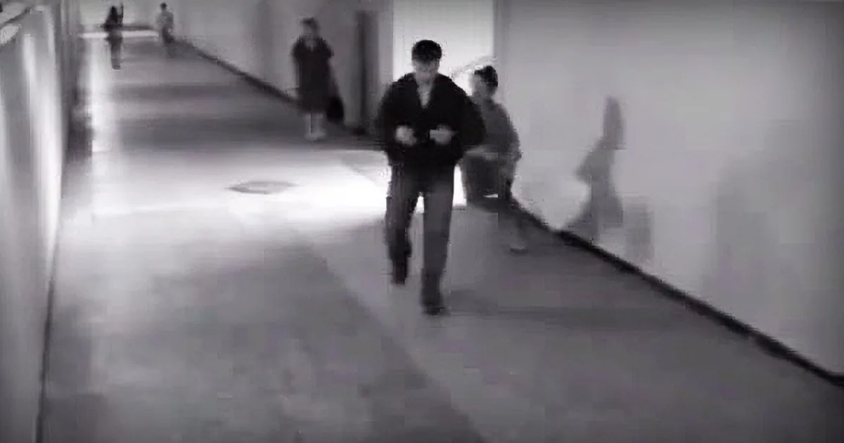A person in a dark jacket walks down a dimly lit hallway, with others in the background.