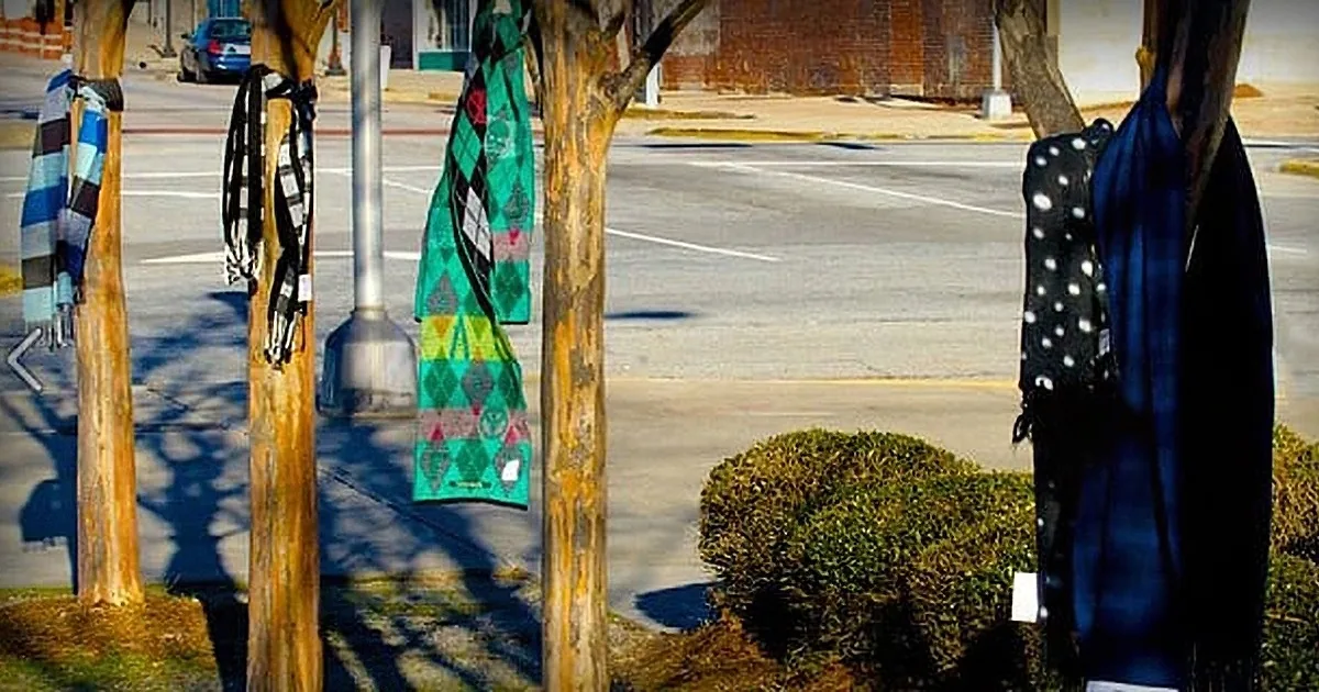 Colorful scarves hang from tree trunks in a city street scene.