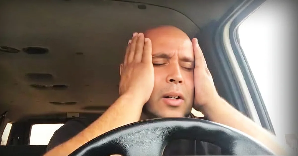 A man in a car covers his face with his hands, appearing distressed or tired.