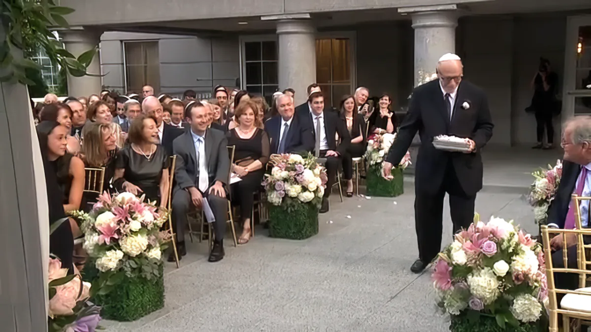 A man in a suit sits at a table with flowers, surrounded by people in a formal setting.
