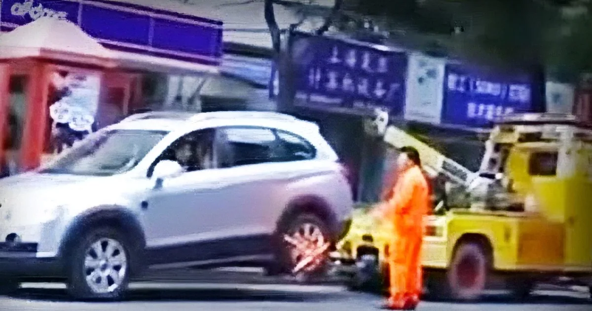 A tow truck is lifting a white car, with workers in orange uniforms assisting, in an urban setting.