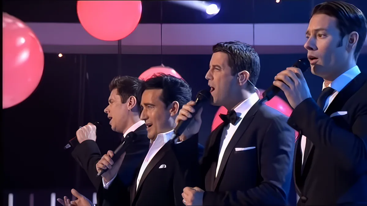 A group of men in tuxedos sing into microphones, with red balloons and stage lights in the background.