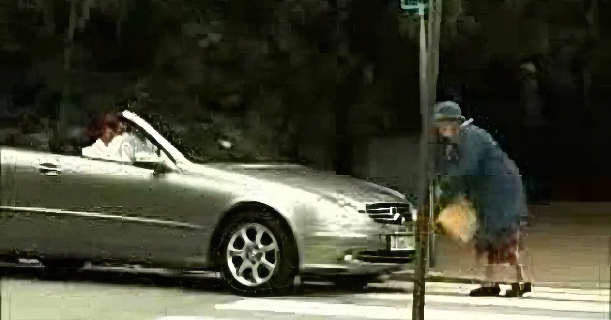 A person in a blue coat walks past a silver car with a person inside on a city street.