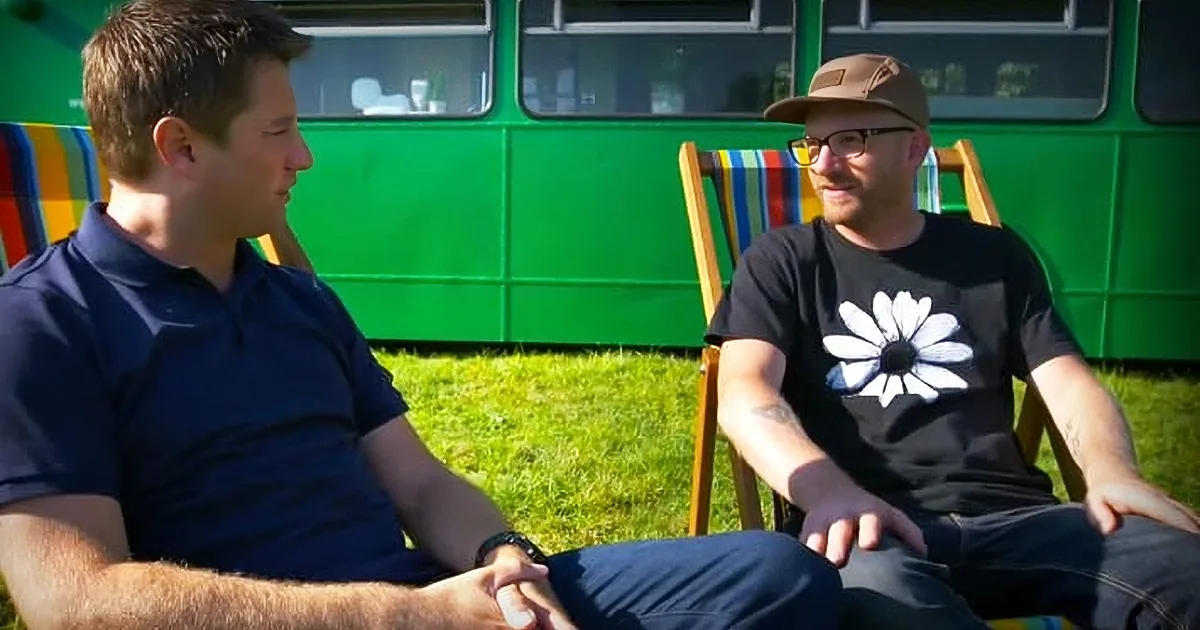 Two men in chairs, one in a blue shirt, the other in a black shirt with a flower, sitting outdoors in front of a green bus.