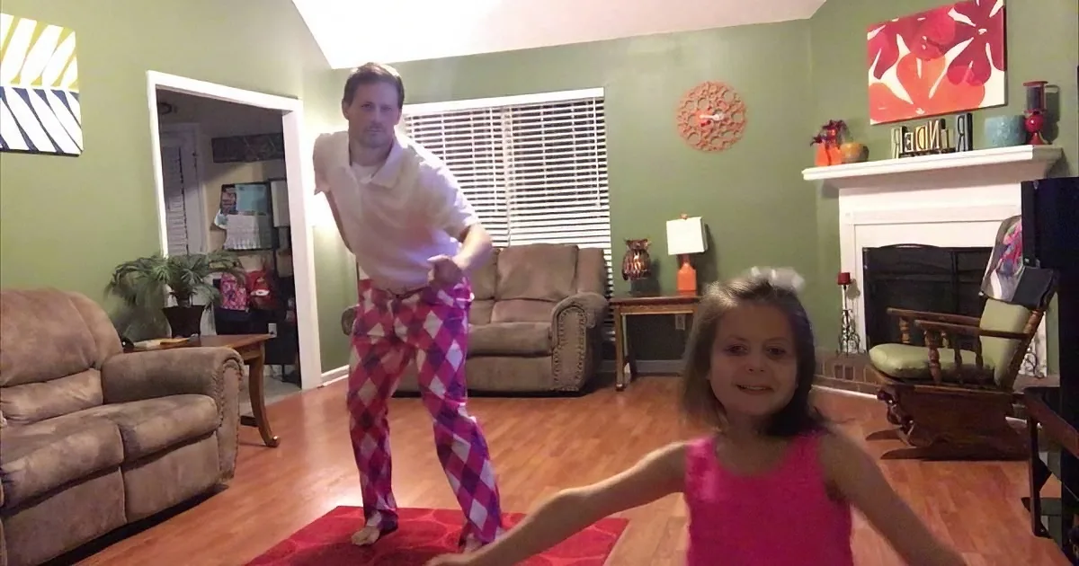 A man in argyle pants and a young girl in a pink dress are dancing in a cozy living room with green walls and a fireplace.