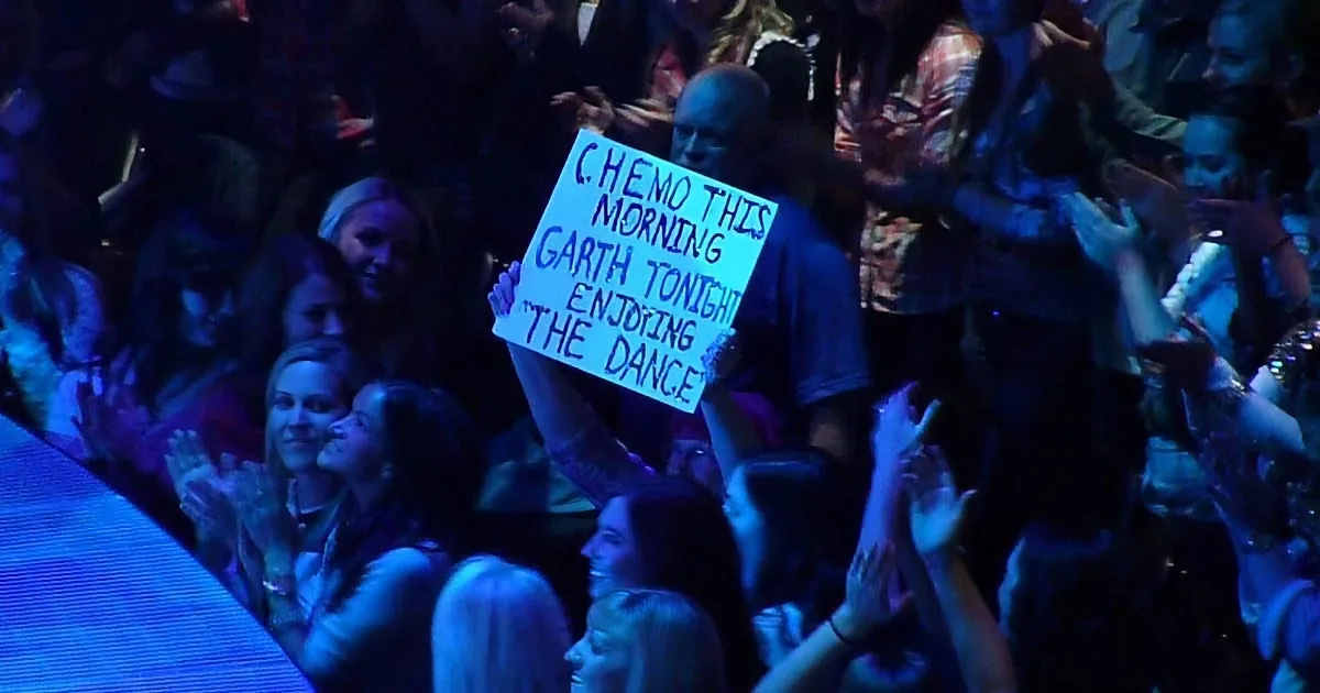 A person holds a sign in a crowd, reading "C. HEMO THIS MORNING, GARTH TONIGHT, ENJOYING THE DANCE."