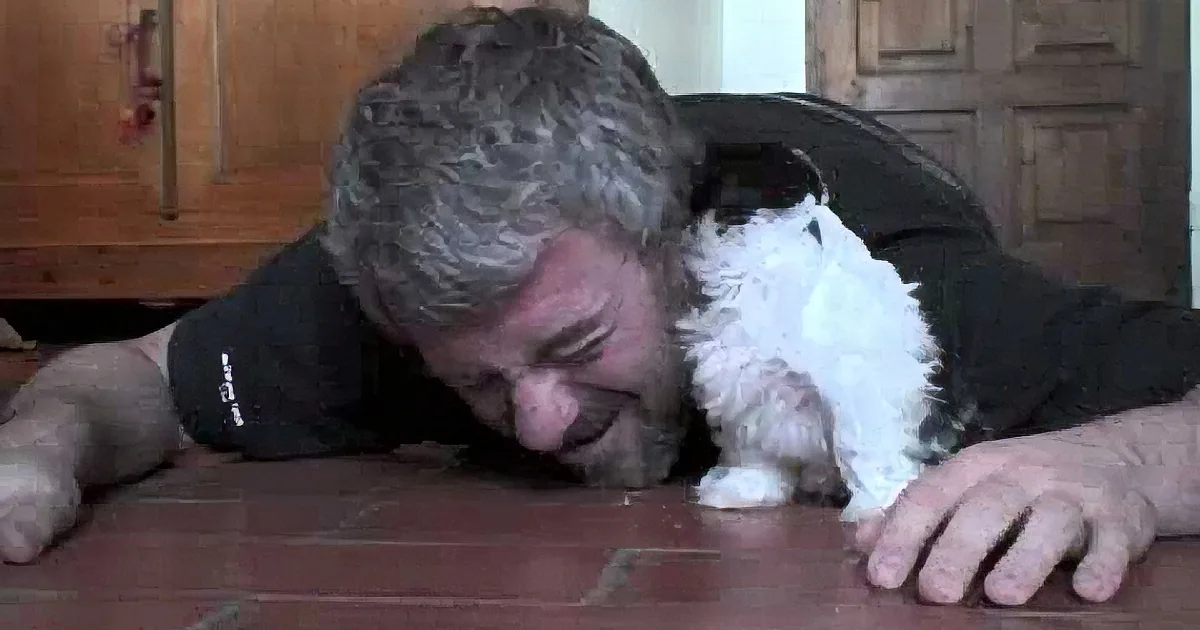A man lies on a tiled floor, embracing a small white dog with black spots.