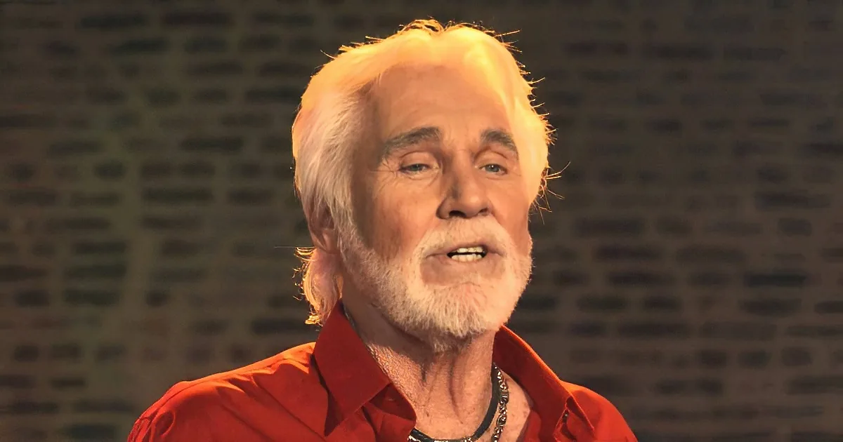 An older man with white hair and a beard, wearing a red shirt, stands against a textured background.