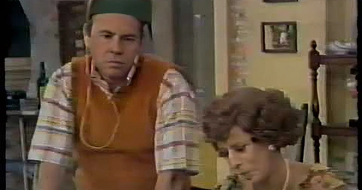 A man in a striped shirt and orange vest stands in a room, looking concerned, with a woman in the foreground.