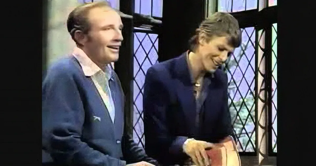 Two men, one in a blue sweater and the other in a dark suit, are seated and smiling in front of a window with a diamond...