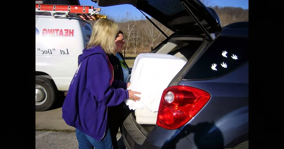 Two women load a white mattress into a car trunk, with a van labeled "Heating" in the background.