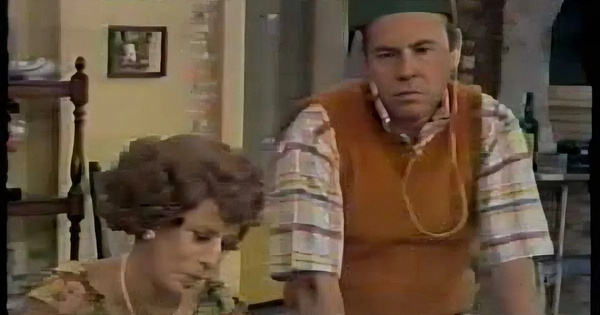 A man in a striped shirt and orange vest stands in a room, looking concerned, with a woman in the foreground.