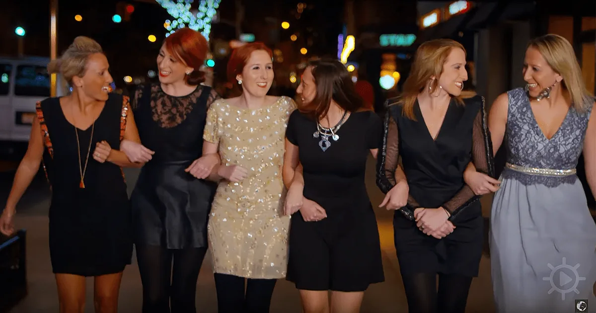 Six women in dresses walk together on a city street at night, smiling and laughing.