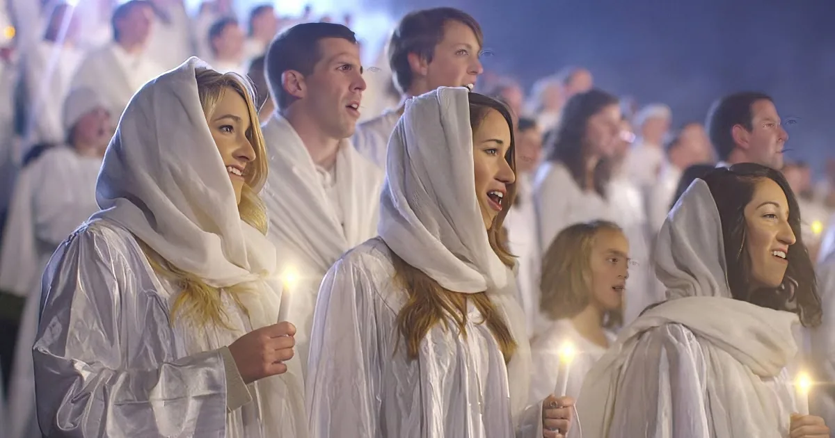 A group of people in white robes and head coverings hold candles, singing in a dimly lit setting.