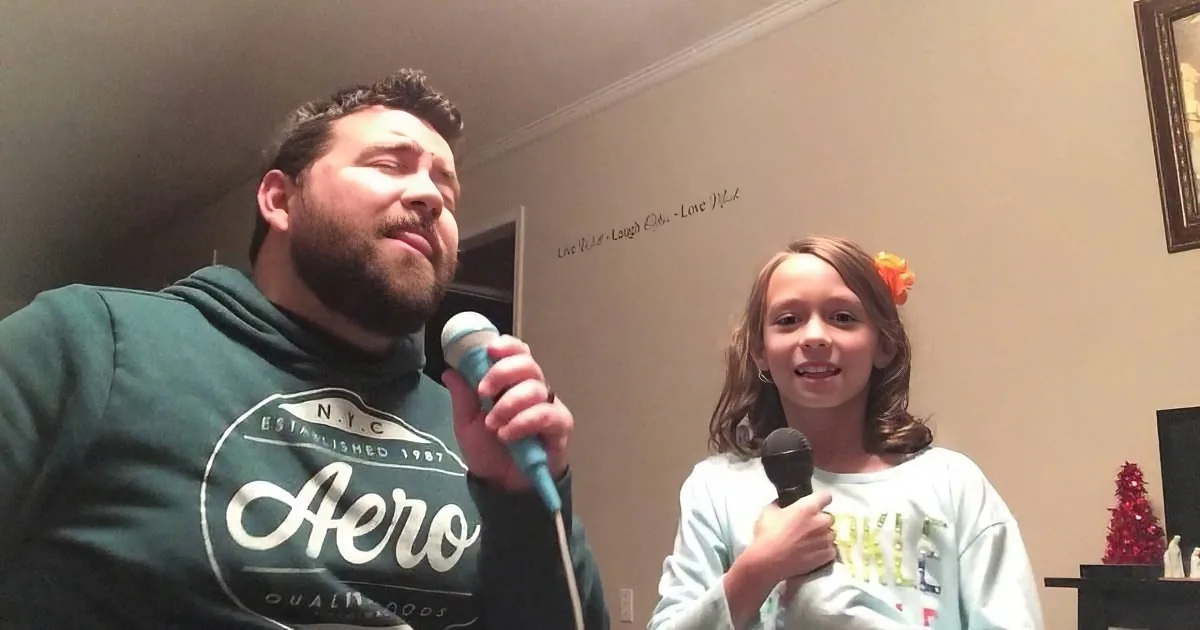 A man and a girl sing into microphones, both wearing hoodies, in a room with a wall decal.