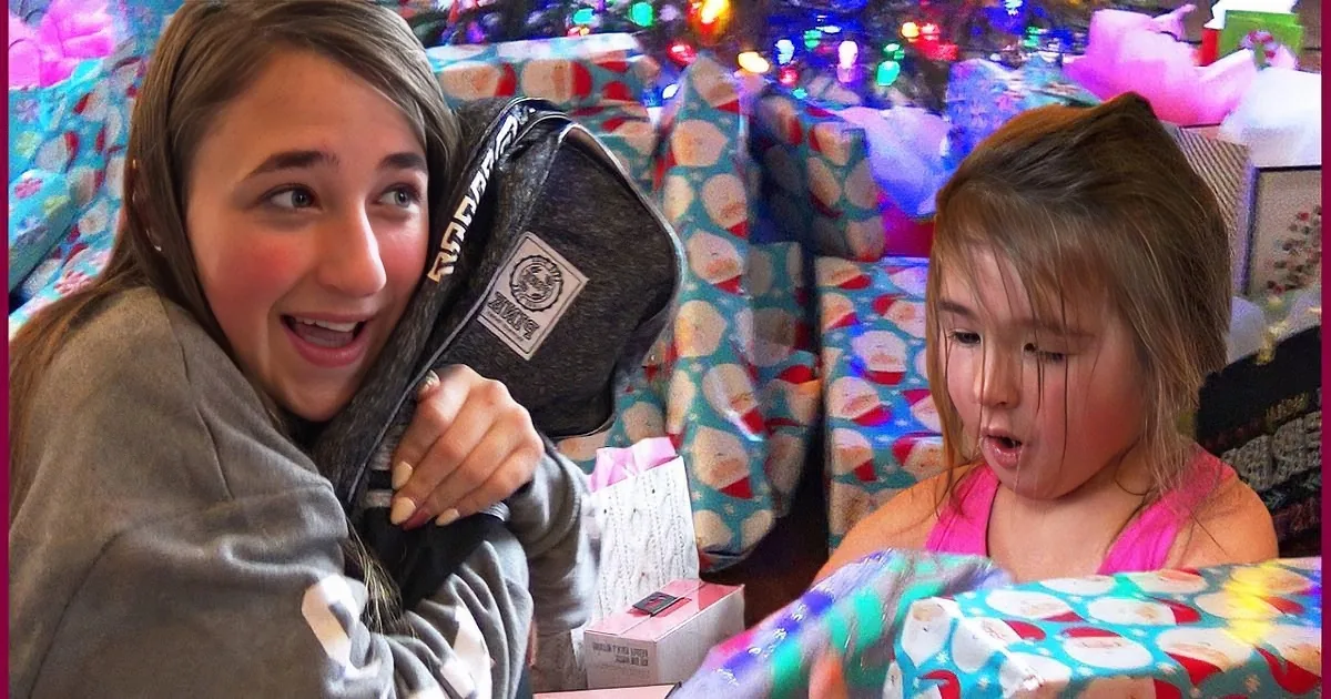 A girl in a gray hoodie holds a black bag, while another girl in a pink top opens a wrapped gift, surrounded by colorful...