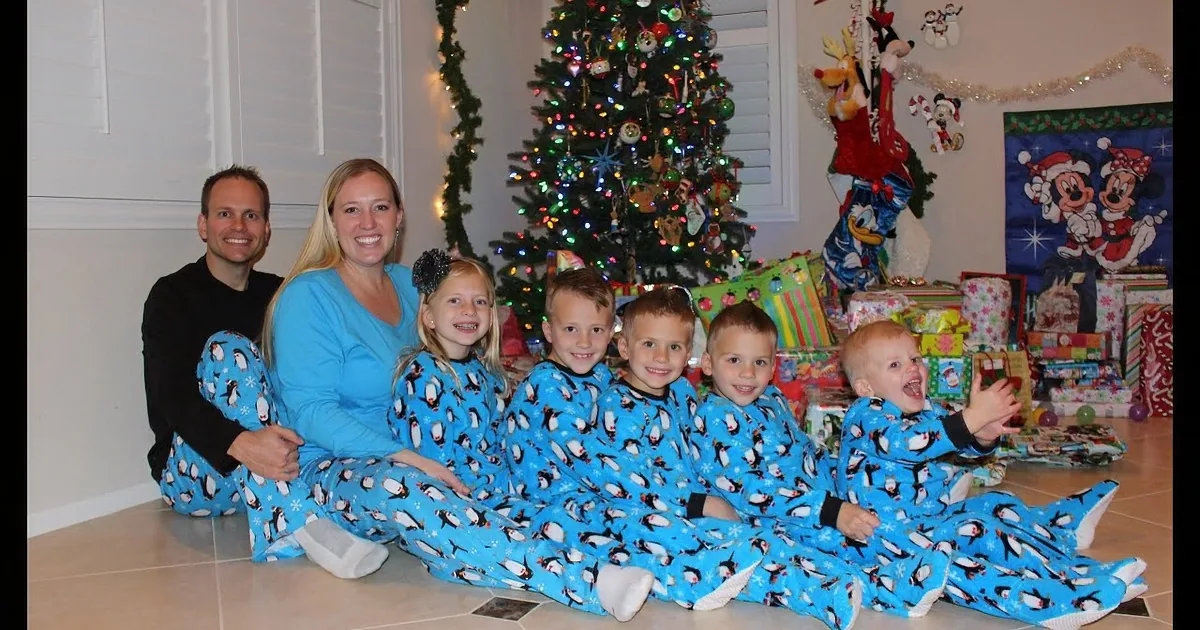 A family in matching blue penguin pajamas poses in front of a Christmas tree with presents and decorations.