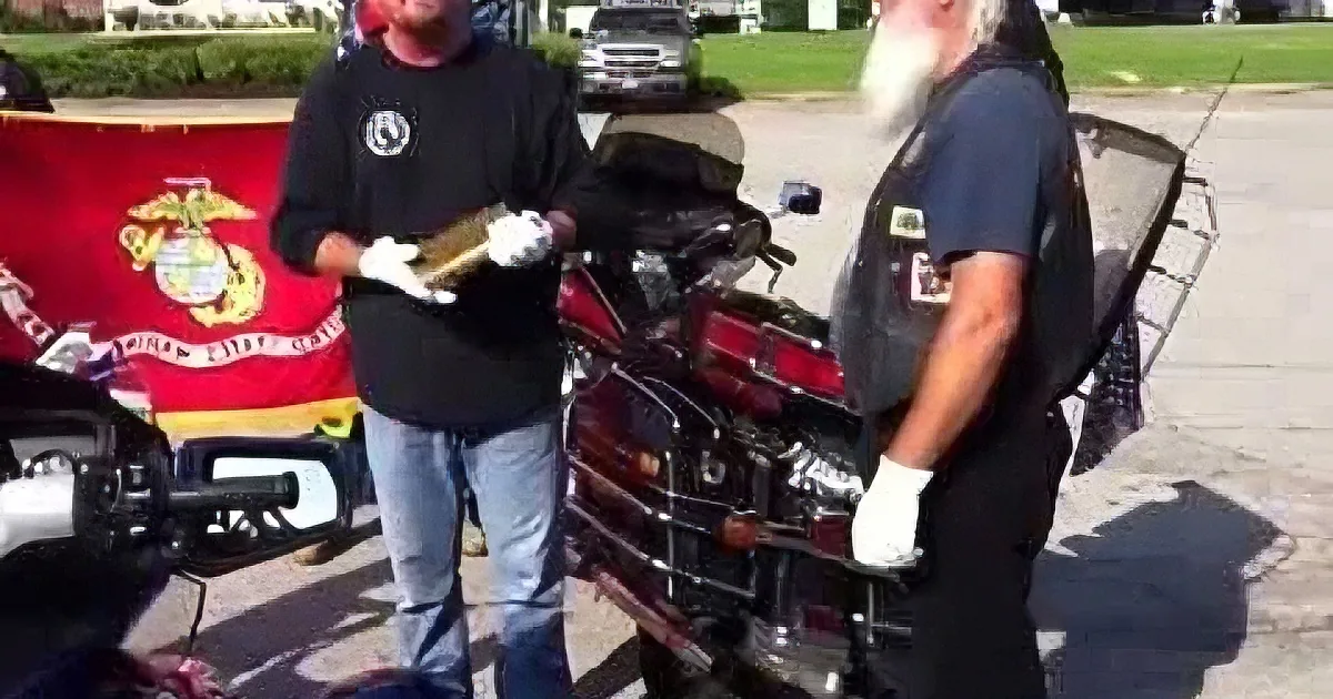 Two men in black shirts and gloves stand beside a motorcycle, one holding a book, with a Marine Corps flag in the background.