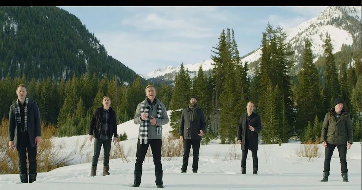 A group of men in winter attire stands in a snowy landscape with evergreen trees and mountains in the background.