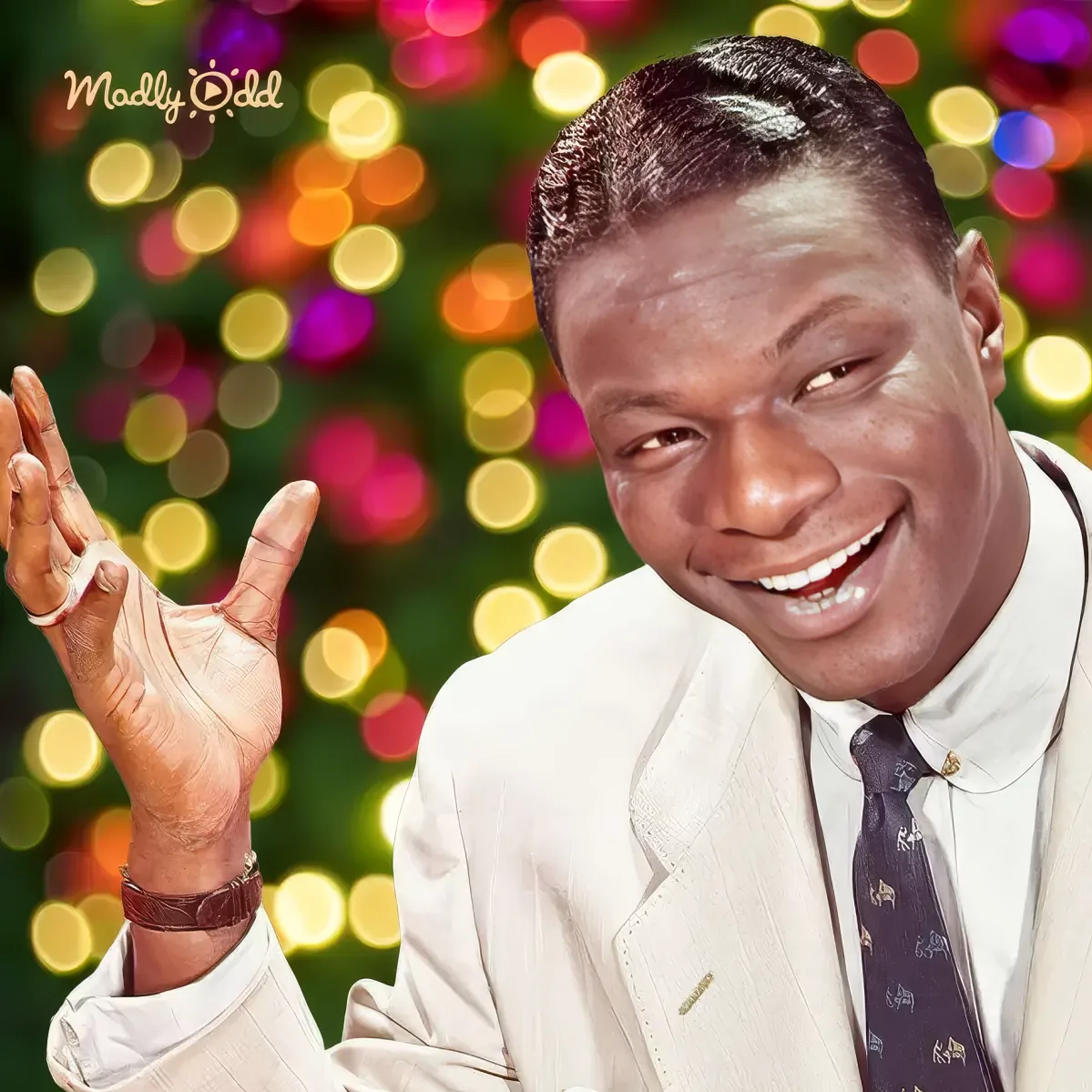 A smiling man in a white suit and tie, raising his hand, with colorful bokeh lights in the background.