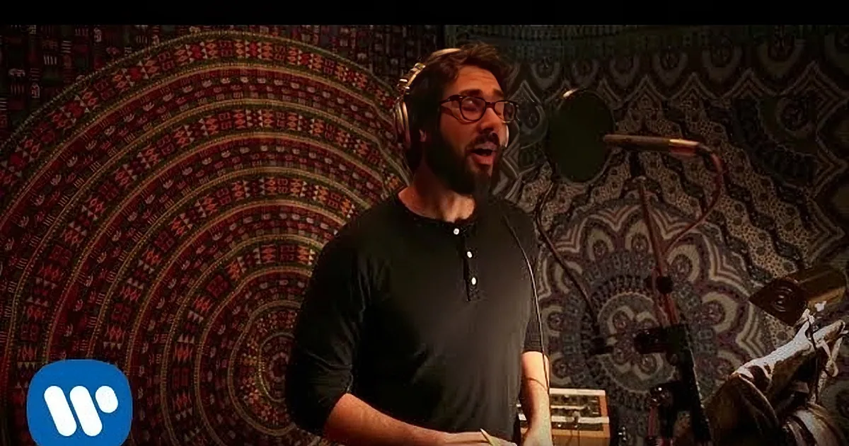 A man wearing headphones and a black shirt sings into a microphone in a recording studio with patterned walls.