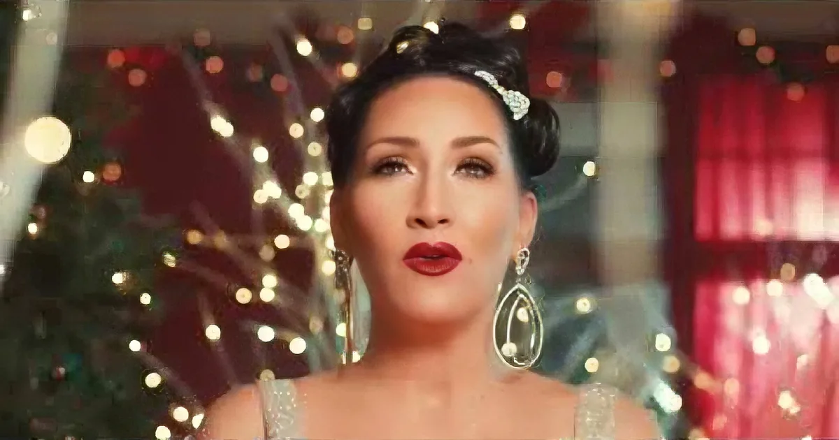 A woman with dark hair and red lipstick, wearing large earrings, stands in front of a Christmas tree with lights.