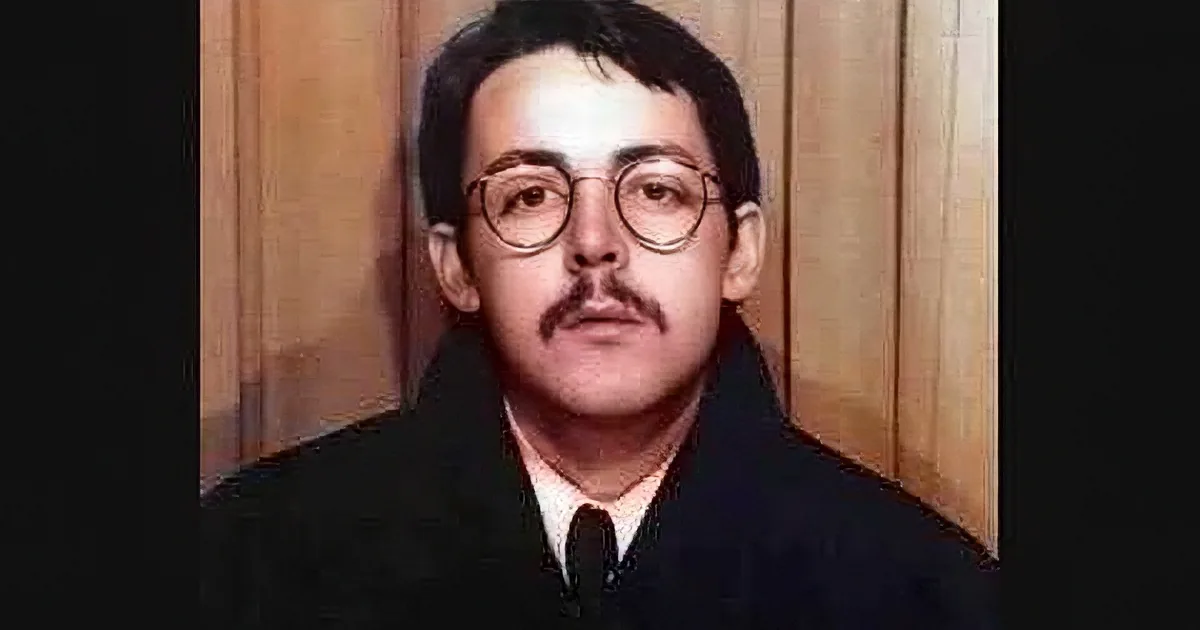 A man with glasses and a mustache, wearing a dark coat and tie, stands against a wooden background.