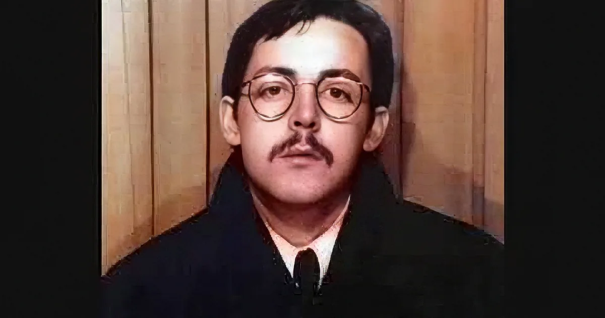 A man with glasses and a mustache, wearing a dark suit and tie, stands against a wooden background.