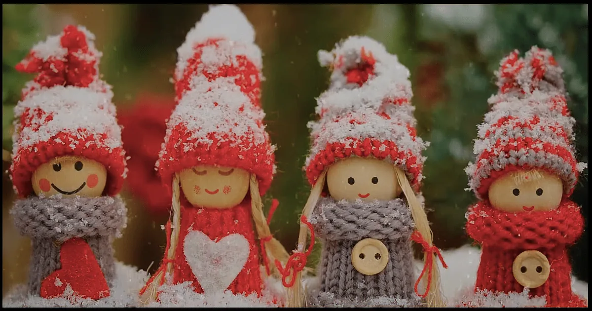 Four snow-covered wooden dolls in red and gray outfits stand in a snowy scene.