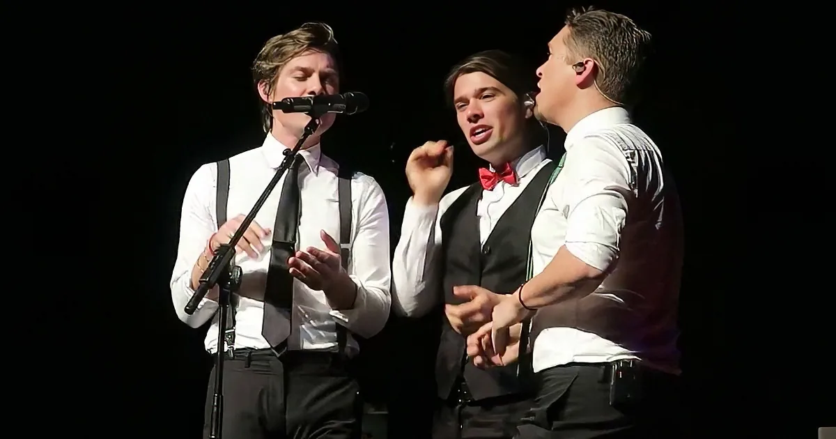 Three men in formal attire sing on stage, one holding a microphone, another in a red bow tie, and the third in a vest.