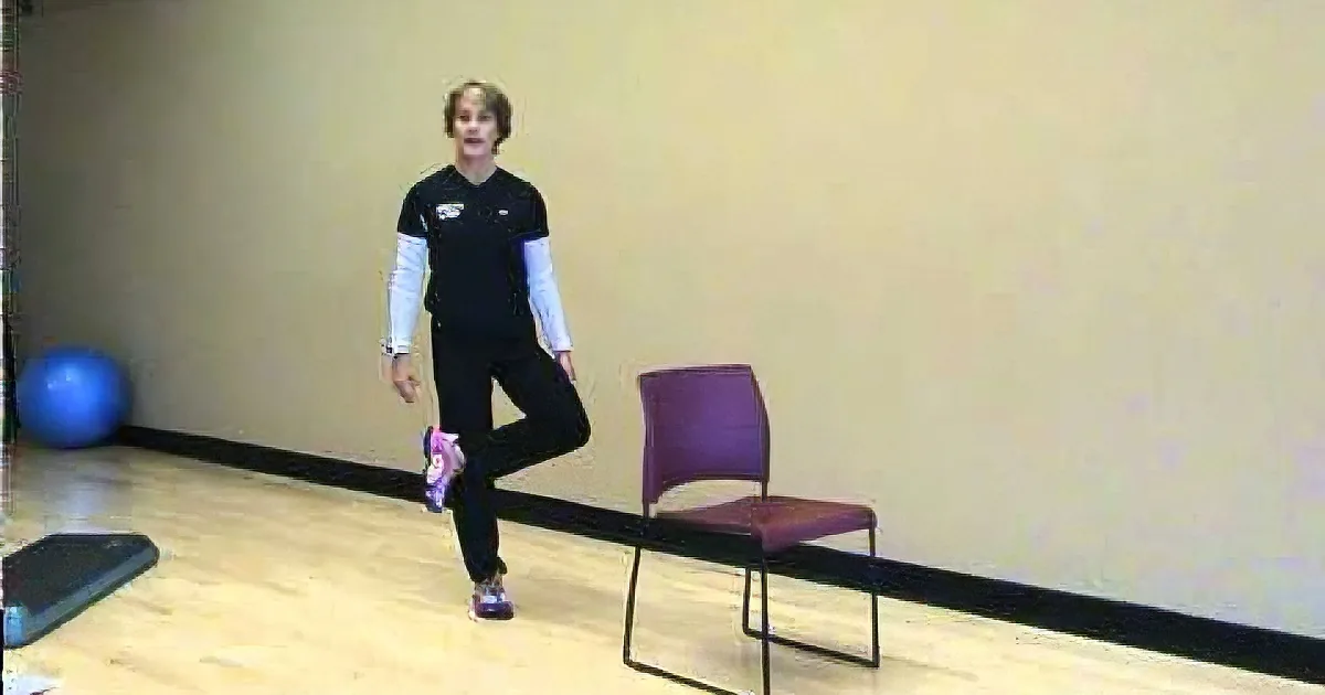A person in a black outfit stands in a balance pose, one leg extended, in a room with a chair and exercise ball.