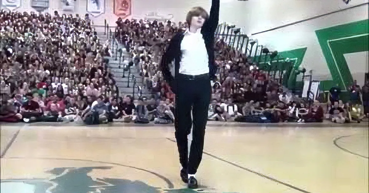 A person in a white shirt and black pants raises one arm on a basketball court, with a large crowd in the background.