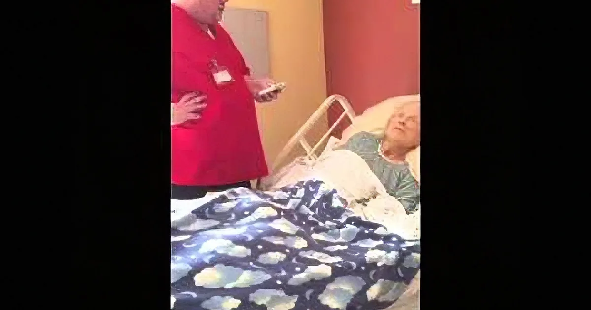 A nurse in red scrubs stands beside a patient in a hospital bed, holding a device.