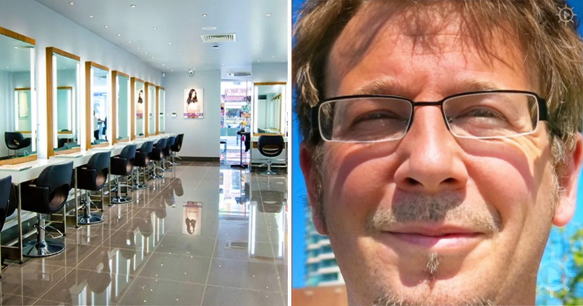 A man with glasses and a beard smiles, next to a salon with multiple mirrors and chairs.