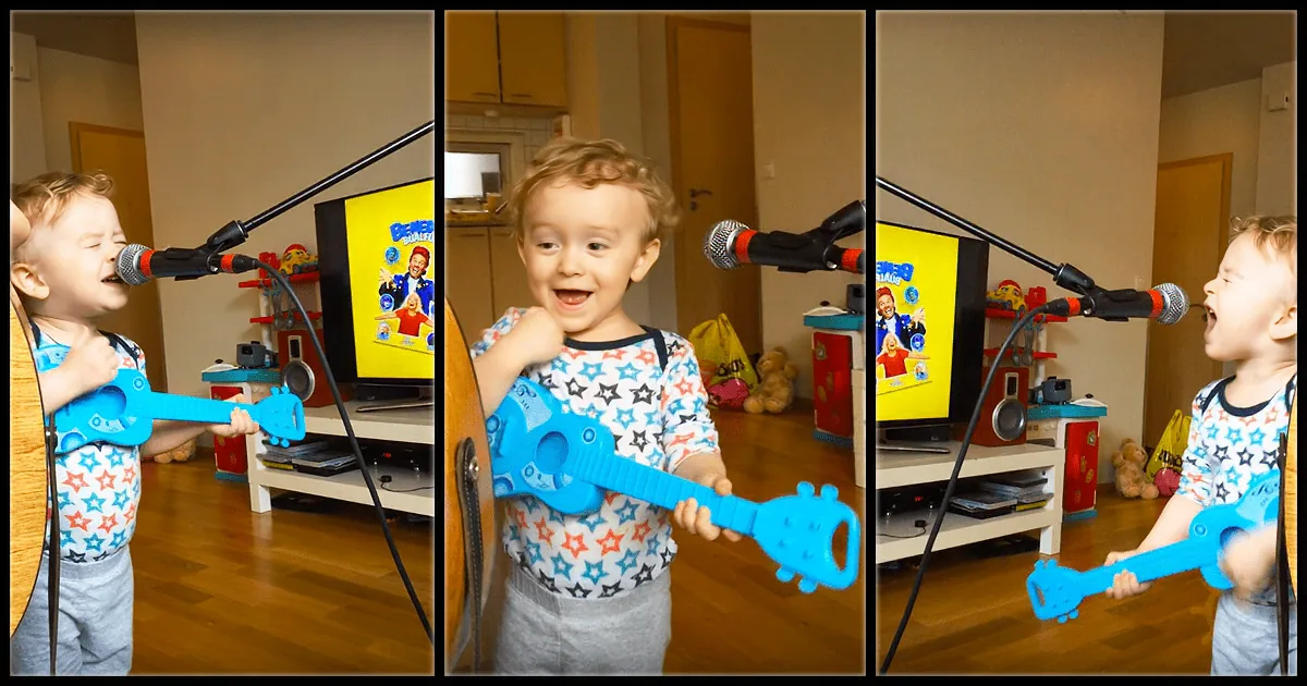 A child plays a toy guitar and sings into a microphone in a room with a TV and toys.