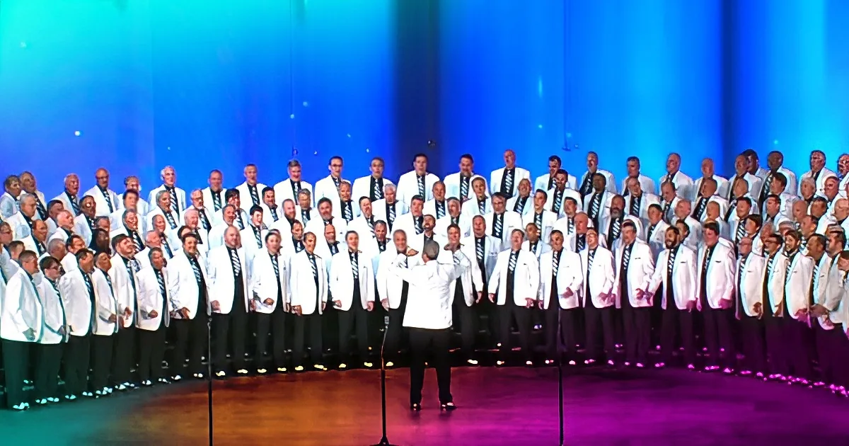 A conductor leads a large choir in white jackets on a stage with colorful lighting.
