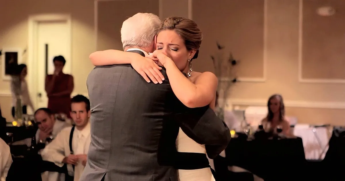 A woman in a white dress hugs a man in a suit, both appearing emotional, with guests in the background.