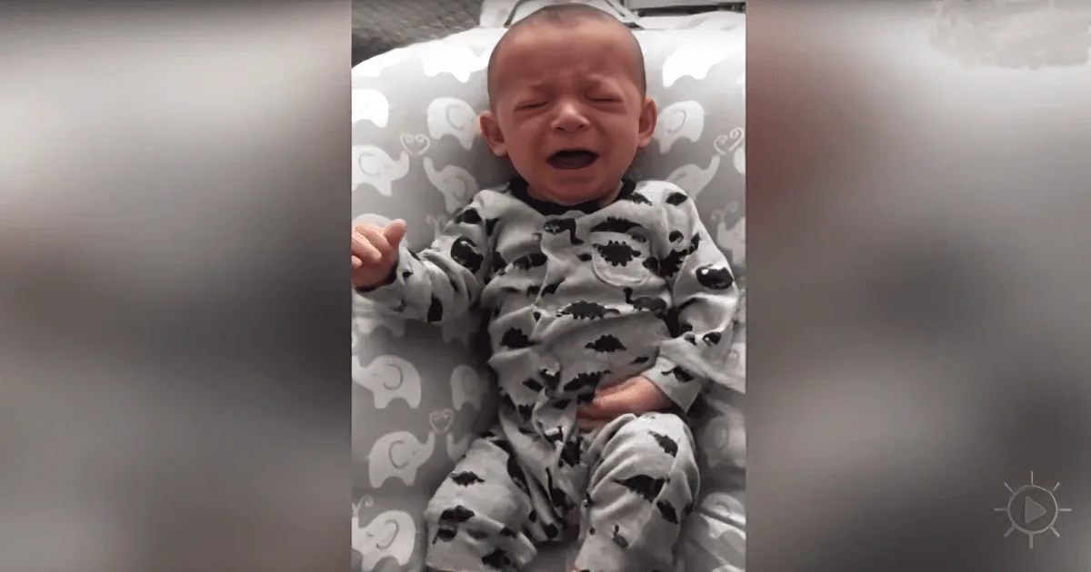 A baby in a grey dinosaur onesie is lying on a patterned surface, appearing to cry.
