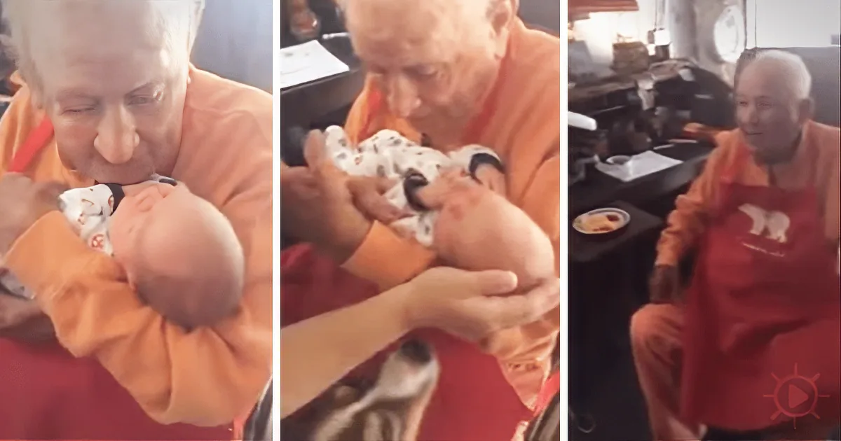 An elderly man in orange holds and gently kisses a baby wrapped in a white blanket with black patterns.