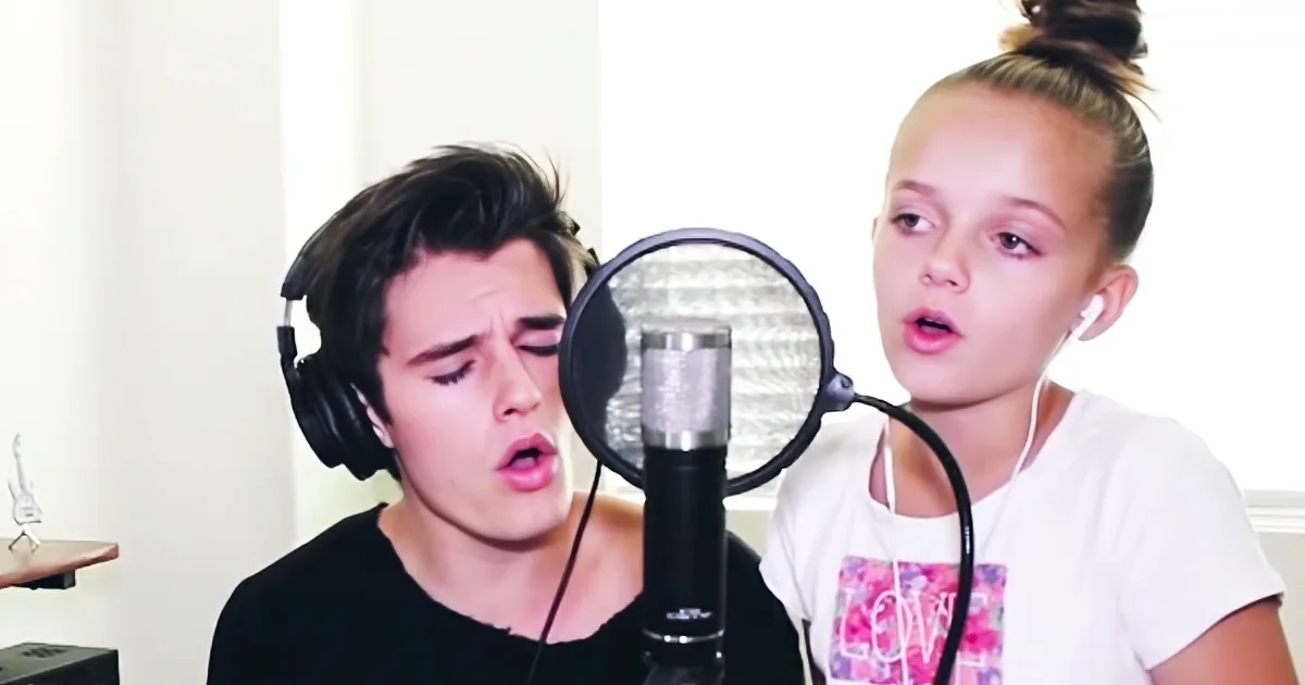 A young man and girl sing into microphones, both wearing headphones, in a recording setup.